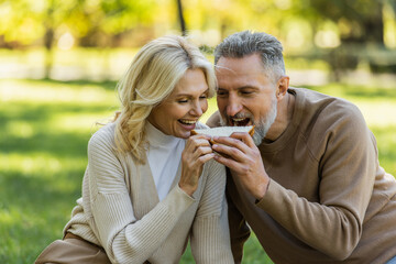 cheerful middle aged couple eating one sandwich from both sides and smiling during picnic in park.