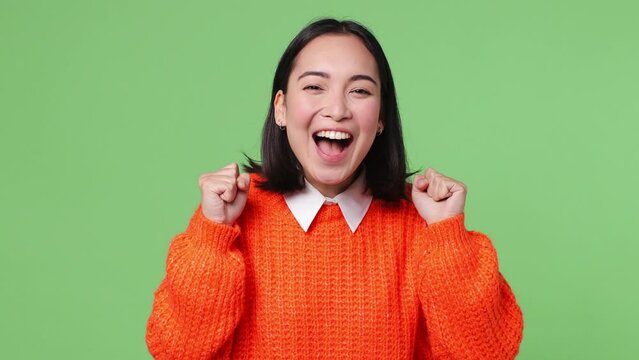 Excited happy young woman of Asian ethnicity 20s she wear orange sweater doing winner gesture celebrate clench fists say yes isolated on plain pastel light green color wall background studio portrait