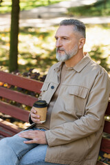 Mature man in spring outfit holding coffee to go while sitting on bench in park.