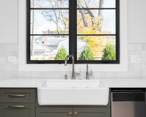 A sink detail shot with an apron sink, green cabinets, black window frame, and a tiled backsplash.