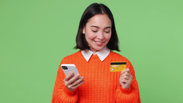 Young woman of Asian ethnicity she wear orange sweater use mobile cell phone app hold in hand credit bank card doing online shopping order to home isolated on plain pastel light green color background