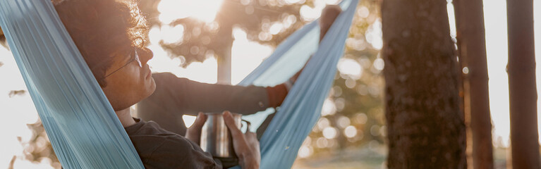 Handsome young man in glasses lying in hammock and relaxing with coffee. Forest travel.