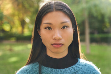 Portrait of beautiful young Asian girl with serious countenance looking at camera outdoors. Front view of earnest Chinese girl. Confident people posing in the park for photo.  - obrazy, fototapety, plakaty