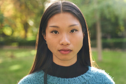 Portrait of beautiful young Asian girl with serious countenance looking at camera outdoors. Front view of earnest Chinese girl. Confident people posing in the park for photo. 