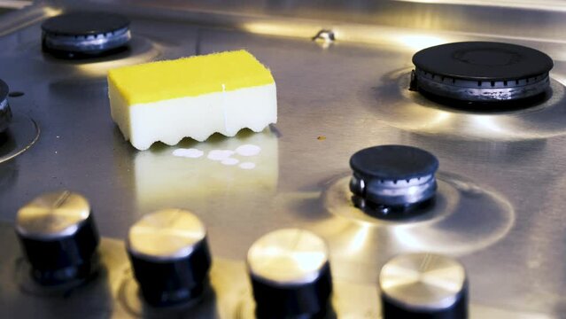 A man applies washing-up liquid to a stainless steel gas surface and cleans it with a kitchen sponge in a protective glove.