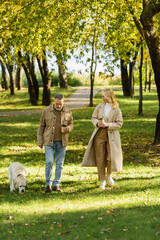 cheerful middle aged woman holding coffee to go and looking at husband walking out with labrador in...