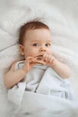 A cute funny little baby with a wood teether is resting in bed after bathing, covered with a towel, laughing happily. The concept of a baby bath. Emotion series. 