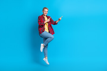 Full length photo of optimistic energetic woman wear checkered shirt jeans directing empty space jump isolated on blue color background