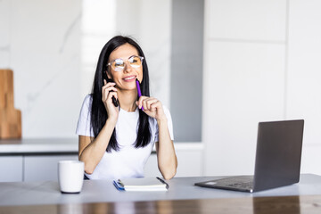 Portrait of a young woman talking the phone and working on laptop in kitchen