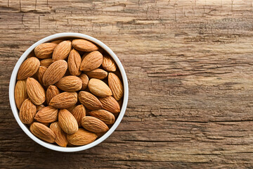 Raw shelled almond seeds in white bowl, photographed overhead on wood with copy space on the side (Selective Focus, Focus on the top almonds)