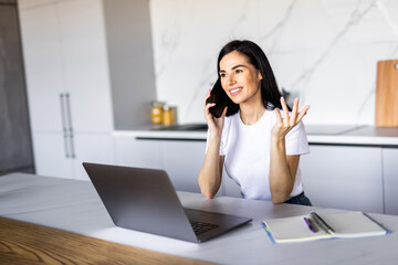 Young woman sits at the kitchen table using a laptop and talking on a phone