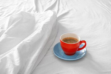 A red cup with a blue saucer with coffee stands on a bed with a blanket
