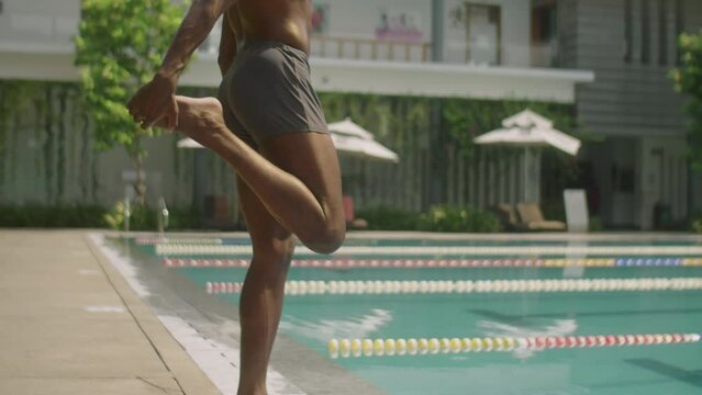 Tilt up shot of professional African American swimmer in cap and trunks standing at edge of outdoor pool and stretching quads before swimming training