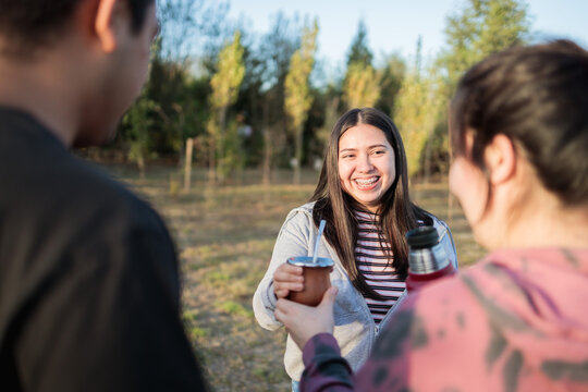  Group of smiling friends drinking yerba mate using a thermo with hot water in the countryside at sunset.