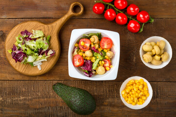 Vegetable salad with champignon corn and cherry tomato with avocado in a white plate on a wooden table next to all the ingredients.