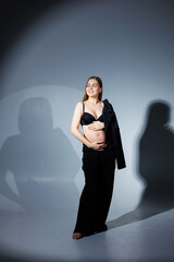 Fototapeta premium Elegant pregnant woman. Beautiful pregnant girl with long hair in black suit on gray background, concept of happy pregnancy and family.