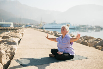 Fototapeta premium Mature woman with dreadlocks working out doing yoga exercises on sea beach copy space - wellness well-being and active elderly age concept