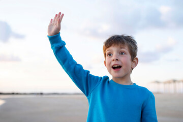 Surprised emotional child waving smiling, beach, sky with clouds in the background. Day light. Concept of travel, vacation, childhood, greeting. Caucasian boy 6 years old.