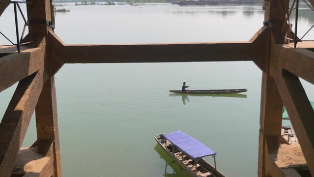 A long pirogue passes under a concrete bridge on a lake in Laos