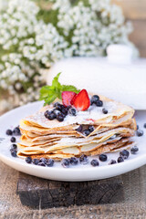 Pancakes with blueberries and mint leaf on top. A bunch of big homemade pancakes with forest fruits. A pile of flat thin cake in the background on a white plate.,wooden background