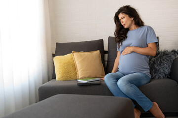 Beautiful 9 month pregnant woman sitting on the sofa at home having back pains caused by carrying a baby