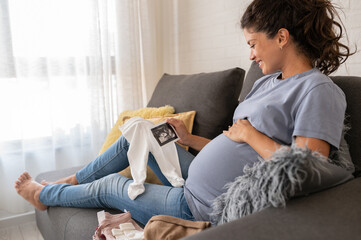 Beautiful pregnant woman looking a cute baby clothes and ultrasound picture at home. Anticipation of a child birth. - obrazy, fototapety, plakaty
