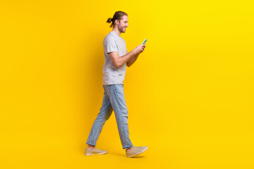 Full size photo of handsome cheerful man gray t-shirt denim pants go empty space look at smartphone isolated on yellow color background