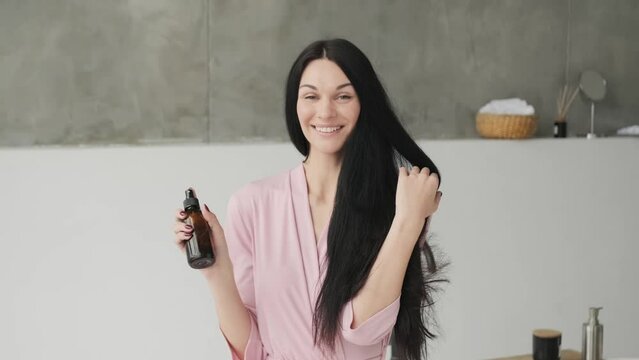Hair Care. happy satisfied woman holding her long healthy black hair and showing at camera Spray shampoo or conditioner, female using natural paraben free hair-care beauty products