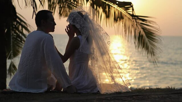 Married couple enjoys atmosphere of sunset against sun-path on ocean. Couple kisses under palm branch on beach after wedding ceremony