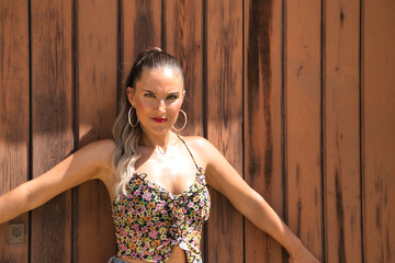 Fototapeta premium Young and beautiful woman, with ponytail, wearing a floral top and jeans, serious and angry with open arms leaning on a wooden door. Concept beauty, seriousness, anger, reproach.