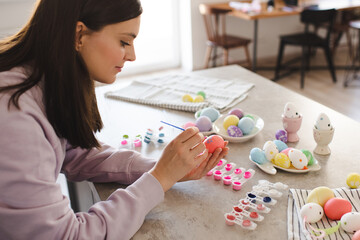 Woman painting colorful Easter eggs with brush over kitchen table with bunny shape plates and dye at home. Holiday season. Springtime.