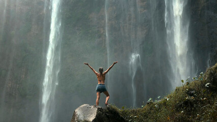 A young woman is facing the waterfall with her hands up. No face.