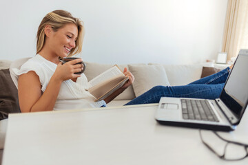 Young girl reading a book and drinking coffee, while holding her feet on the table. Books on table with woman and computer laptop at the background.