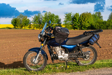 Fototapeta premium A motorcycle stands on the road against the background of a dug-up agricultural field on a sunny day. A motorcycle trip through nature. Motorcycle on a rainbow background.