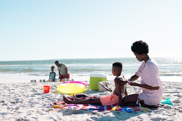 African american woman applying sunscreen on son's body while sitting on beach against sky
