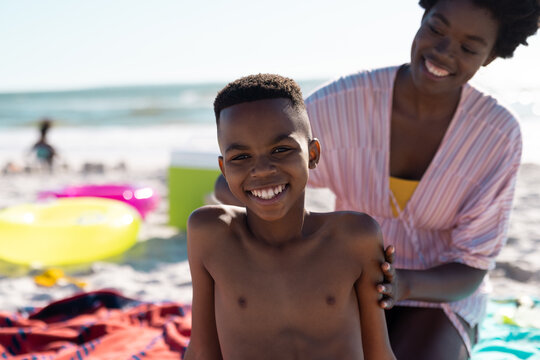 African american mother applying sunscreen on son's back while sitting at beach against sky