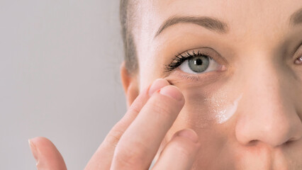 Beauty young smiling woman applying cosmetic cream under her eye on a white studio background
