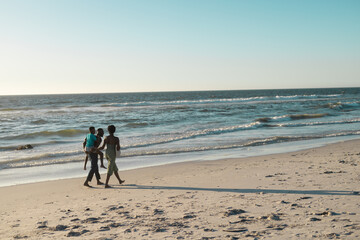 African american man piggybacking son and walking with wife at beach by beautiful sea against sky