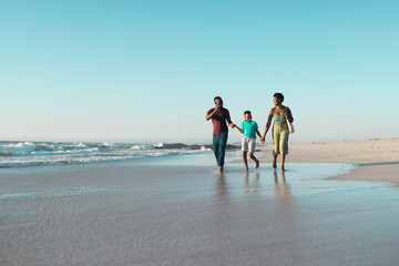 Fototapeta na wymiar Cheerful african american boy holding father and mother's hands and running in sea against sky