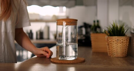 Anonymous female hand on countertop glass jug with water in kitchen. Unrecognizable woman in white tshirt stands near jar with cork cap at home.