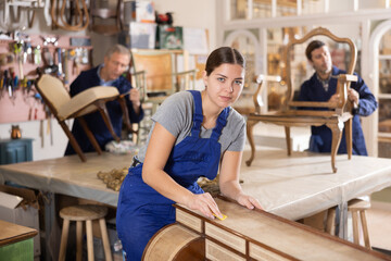 Skilled female furniture repair specialist in uniform restoring Victorian dresser during workday in workshop