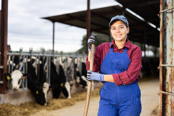 Hardworking young woman farmer in overalls posing with rake and smiling at camera while working on cow farm © JackF