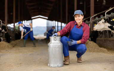 Portrait of young European woman dairy farmworker in uniform carrying metal milk can in cowshed