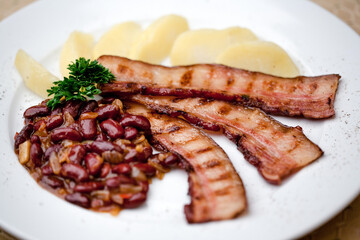 grilled bacon, slices of bacon on a plate with beans and boiled potatoes, lunch in the pub