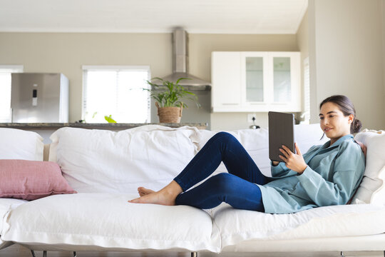 Side view of caucasian young woman using digital tablet while relaxing on sofa at home, copy space