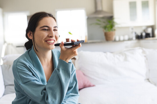 Smiling caucasian young woman talking on speaker over smart phone while sitting on sofa at home