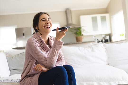 Cheerful caucasian young woman talking on speaker over mobile phone while sitting on sofa at home