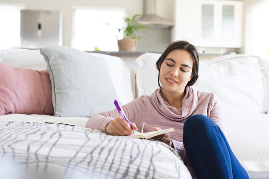 Caucasian young woman writing in notepad while sitting by sofa in living room