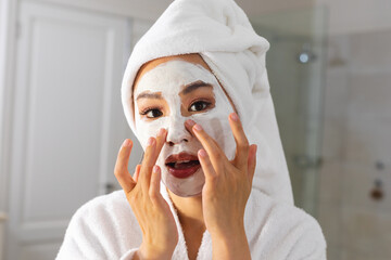 Asian woman wearing bathrobe and towel on head looking in bathroom mirror applying beauty face mask