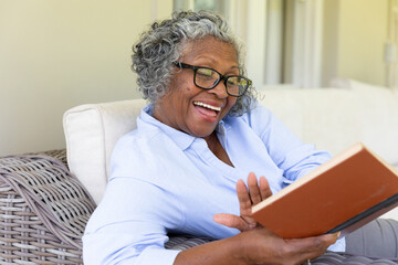 Cheerful african american senior woman laughing and reading book while relaxing on sofa at home